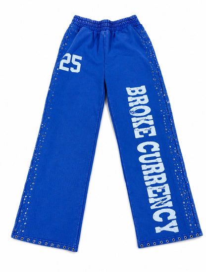 Blue wide-leg sweatpants with white text reading 'BROKE CURRENCY' down one leg, a white '25' on the other leg, and silver metal studs along the outer seams, shown on a plain white background.