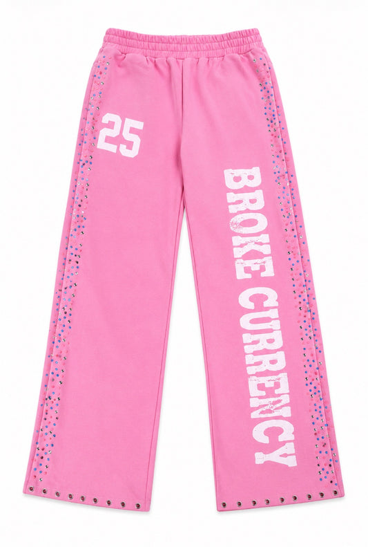 Pink sweatpants with 'BROKE CURRENCY' printed vertically in white on one leg, the number '25' on the other leg, and multicolored studs along the side seams and hem.