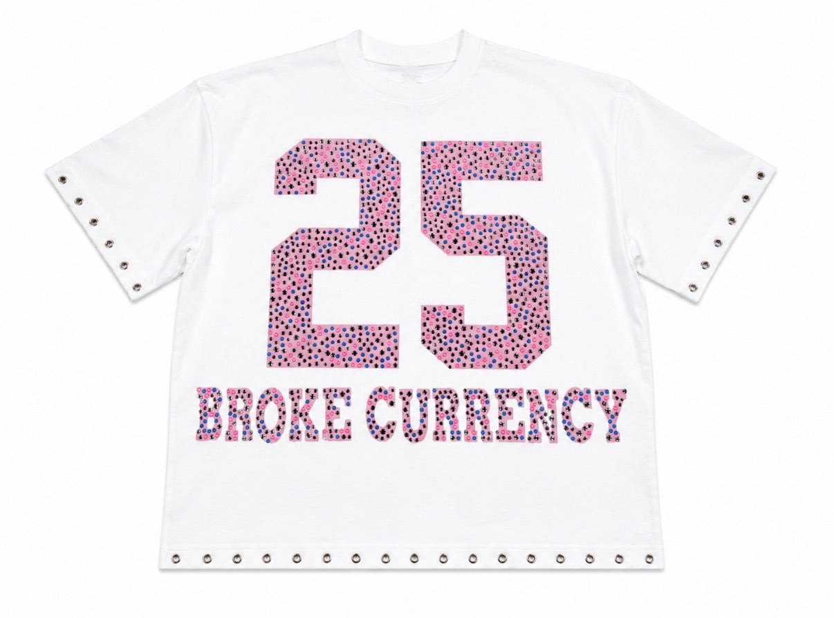 A white short-sleeve T-shirt with decorative silver grommets on the sleeve cuffs and bottom hem, featuring a large pink and purple patterned '25' and 'BROKE CURRENCY' text on the front.